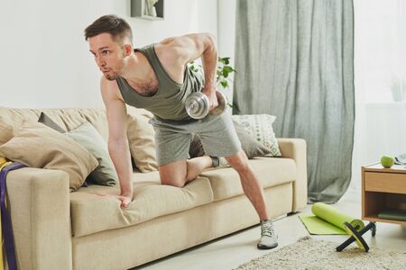 Young sportsman in activewear leaning on couch while doing exercise for armsの写真素材
