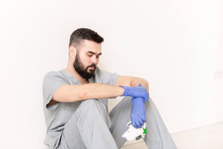 Tired young clinician in protective gloves and grey uniform holding respiratorの写真素材