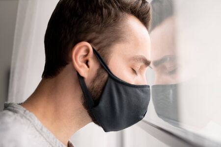 Sad puzzled young man in cloth mask leaning head on window and keeping eyes closed while feeling...