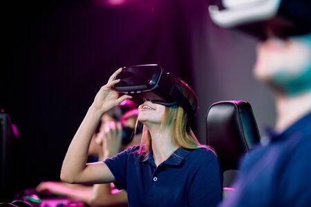 Happy young female video gamer in vr headset sitting among her friends in cybersports gaming club...
