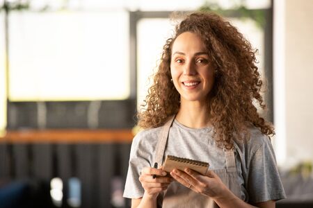Young cheerful waitress with notepad and pen receiving your order in cafeの写真素材