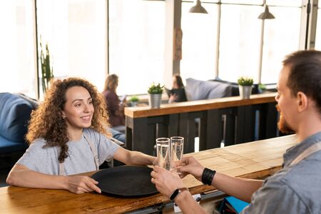 Happy young waitress looking at colleague putting two glasses of water on trayの写真素材