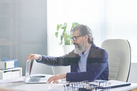 Elegant senior director of business company opening laptop while sitting in armchair by desk and going to get new information in the netの写真素材