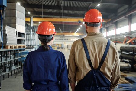 Rear view of industrial workers in orange hardhats contemplating large factory warehouseの写真素材