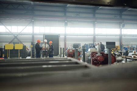 People in hardhats using tablet and discussing engineering project while working at compressor plant