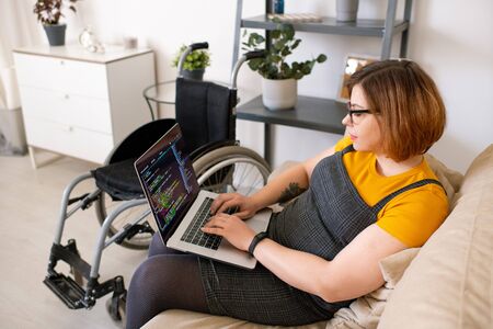 Serious disabled woman in eyeglasses sitting on sofa and using laptop for coding at homeの写真素材