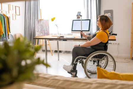 Disabled young woman sitting in wheelchair and using monopod while taking selfie at home