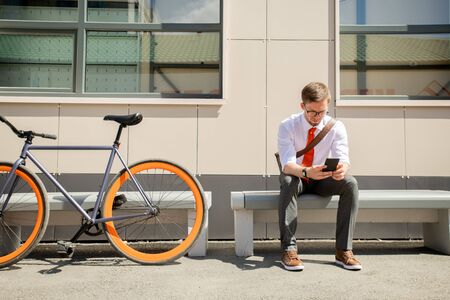 Elegant young man messaging in smartphone while sitting on bench outdoorsの写真素材