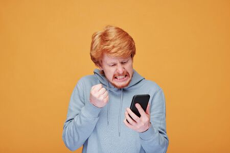Very angry young bearded man in casualwear looking at screen of smartphone