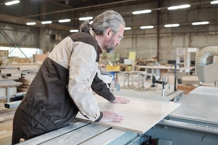 Senior man in workwear bending over wooden board while going to cut it