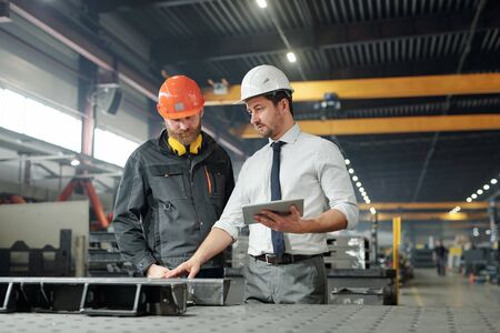 Industrial manager with tablet talking to worker and examining produced metal parts in factory shop