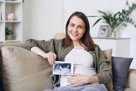 Cheerful future mother with toothy smile showing ultrasonic picture of her baby