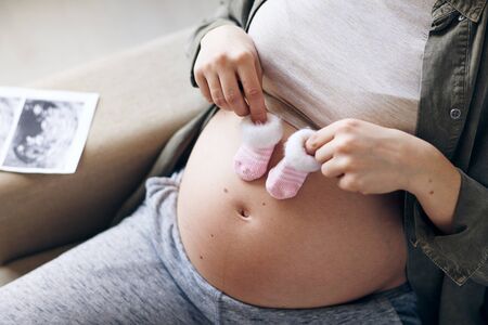 Hands of young pregnant female holding tiny socks for her future baby over belly