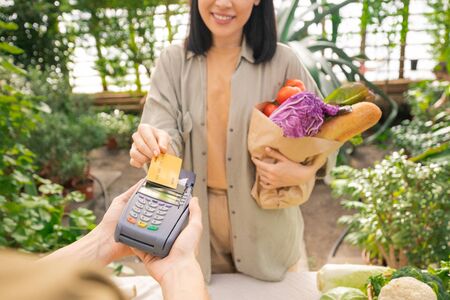 Close-up of smiling lady holding paper bag full of vegetables and paying for organic food with credit card in grocery storeの写真素材