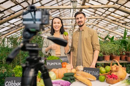Positive young multi-ethnic couple showing apple to camera while shooting video review in greenhouseの写真素材