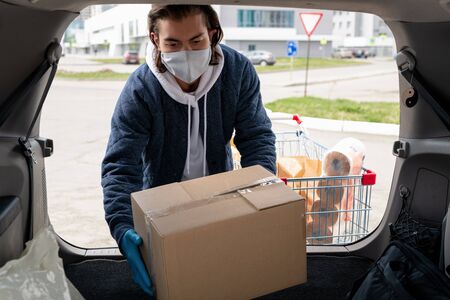 Middle-aged man in cloth mask and jacket standing at car and loading box into trunk after visiting storeの写真素材