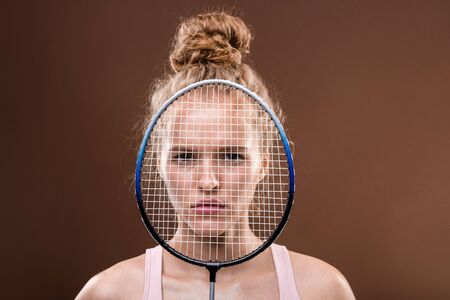 Serious sportswoman with blond hair looking at you from behind tennis racketの写真素材