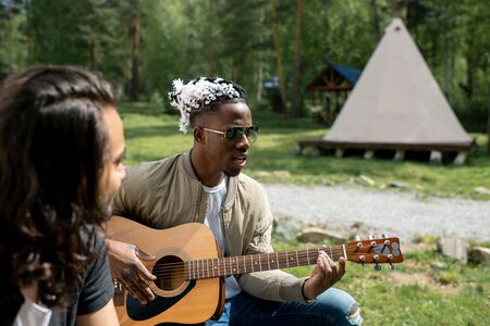 Friends enjoying camping: Afro-American musician in sunglasses playing guitar while singing song with friend at festivalの写真素材