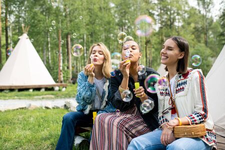 Positive young multi-ethnic women sitting at tent in forest and blowing soap bubblesの写真素材