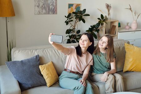Smiling mother sitting on sofa in living room and taking selfie with daughter on smartphoneの写真素材