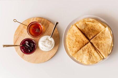 Overview of fresh homemade folded pancakes on plate and small glass bowls with honey, cherry jam and sourcream on wooden boardの写真素材