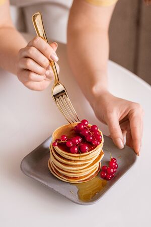 Young woman with fork bending over table while taking fresh red currant from top of pancake stack with honey during breakfastの写真素材