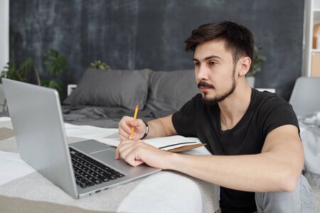 Serious adult student with beard sitting at bed and making notes in diary while watching webinar at homeの写真素材