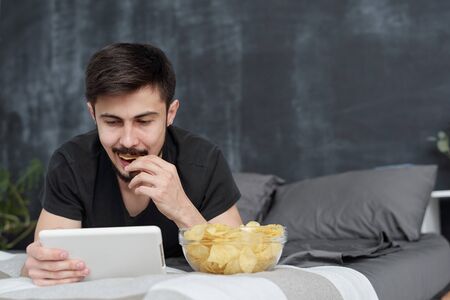 Brunette young guy lying on bed and eating chips while watching video on tablet at homeの写真素材
