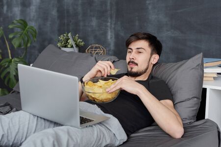Lazy young bearded man lying on bed and eating chips while watching movie on laptop during coronavirus quarantineの写真素材
