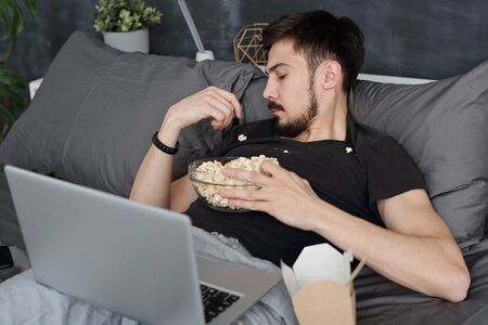 Young man having got popcorn all over him lying with laptop on bed and eating popcorn from tshirtの写真素材