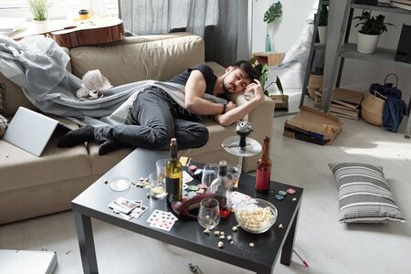 Drunken young man sleeping on sofa among messy stuff in dirty room with alcohol bottle on tableの写真素材