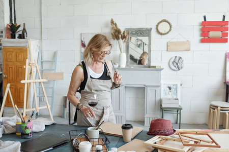 Blond-haired artist in eyeglasses standing at table and drinking wine while thinking about new pictureの写真素材