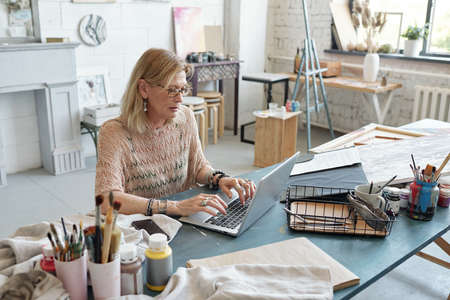 Concentrated mature artist in eyeglasses sitting at table and typing on laptop while buying consumable item for paintingの写真素材