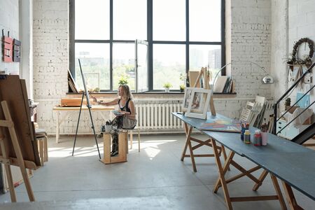 Female artist sitting in front on canvas and painting picture in own spacious studioの写真素材