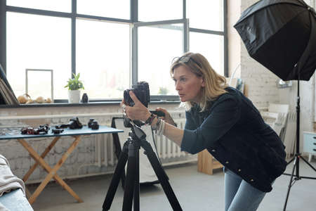 Focused mature woman with eyeglasses on head working with photographic camera in modern photo studio