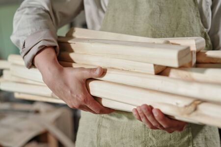 Close-up of unrecognizable carpenter in apron carrying heap of wooden planks to make furnitureの写真素材