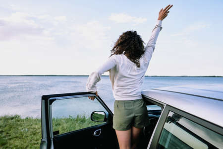 Back view of joyful young woman in casualwear coming out of car and raising her hand while standing...