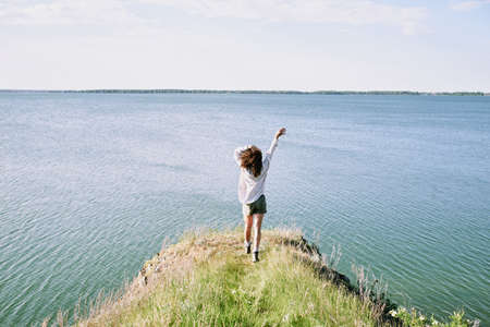 Back view of young ecstatic woman with dark curly hair enjoying freedom and sunny day while moving towards water with her arms raisedの写真素材