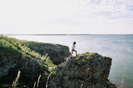 Young female hiker moving along large rock by lake while going towards water on background of seaside, skyline and cloudy skyの写真素材