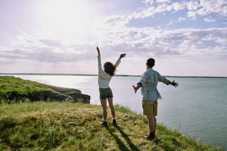 Rear view of young ecstatic couple in casualwear standing on green grass on riverbank and enjoying sunny weekend by seasideの写真素材