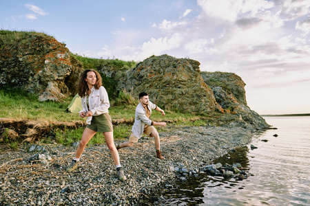 Happy young couple in boots, shorts and shirts throwing stones in water