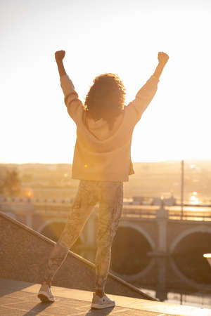 Back view of young ecstatic sportswoman in leggins and hoodie keeping her arms raised while expressing success or gladnessの写真素材