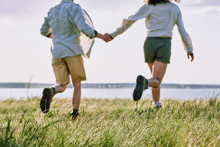 Back view of happy amorous young couple running down green grass towards river or lake while spending weekend in the countryの写真素材