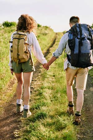 Rear view of two young affectionate hikers with backpacks holding by hands while coming down country...
