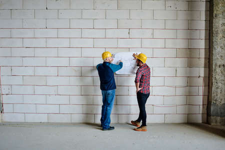 Two professional engineers in hardhats standing by wall and discussing sketchの写真素材