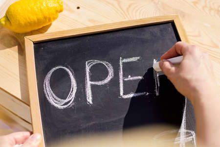 Hand of young seller of cool drinks writing down word open on noticeboard over wooden table with fresh lemon near byの写真素材