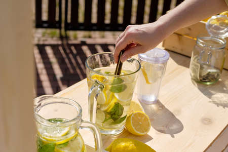 Hand of young woman squeezing lemon slices in glass jug with water while standing by wooden table and making fresh lemonadeの写真素材