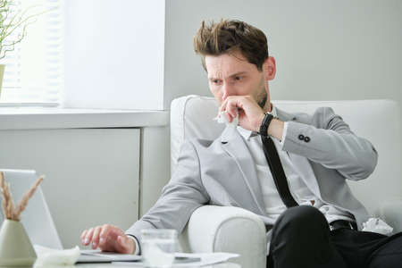 Frowning ill young businessman sitting in armchair and working on project using laptop while having coldの写真素材