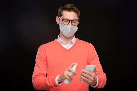 Young man in mask and eyeglasses disinfecting smartphone with sanitizer against black background,...