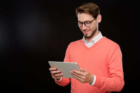 Smiling handsome young bearded man in bright sweater surfing social media on digital tablet against...